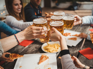 Group of friends toasting with a glass of beer while eating pizza - Millennials have fun together - Day of happiness between young men and women