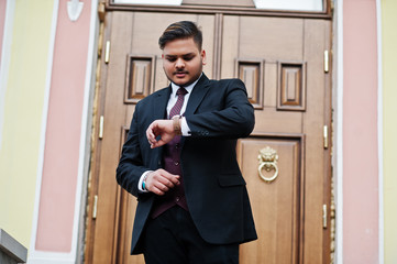 Stylish indian businessman in formal wear standing against door in business center and look at his watches on hand.