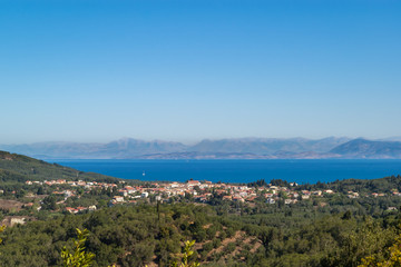 Beautiful panoramic view of the Ionian island of Corfu with olive trees, a small village, the blue waters of the Ionian Sea and high mountains in the background. Half landscape, half blue sky.