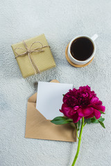 Morning coffee mug for breakfast, present box or gift, empty note, card and pink peony flowers on light stone table. Top view in flat lay style, copy space