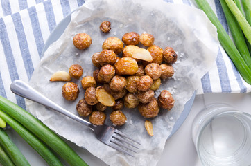 Top view of roasted new potatoes on the baking paper and on the plate,green onion,fork, glass of water, striped napkin on the white table