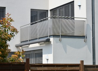 Moderner Balkon mit elektrischer Markise, Balkonlampe, Edelstahl-Geländer, Jalousien und in die Außenwand integriertes Regenfallrohr an einer Neubau-Hausfront