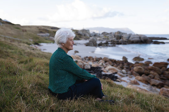 Thoughtful active senior woman sitting on grass at beach