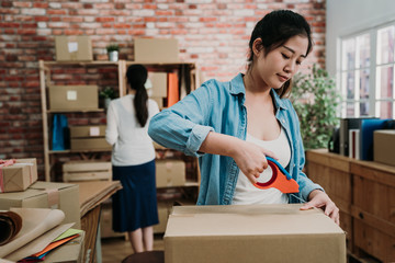 front view of young startup small business woman owner packing cardboard at workplace using tape....