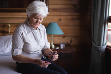 Active senior woman with glucometer checking blood sugar level on bed in bedroom at home