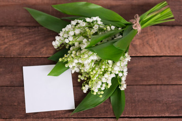 bouquet of delicate white lilies of the valley on a brown wooden background with a white paper sheet for text