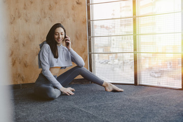 Young attractive fitness girl sitting on the floor near the window on the background of a wooden wall, resting on yoga classes and talking on phone