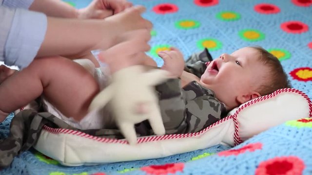 Mom removes clothes from a baby. Baby is playing with a soft toy while lying on a bed. Clothes are being removed from him.