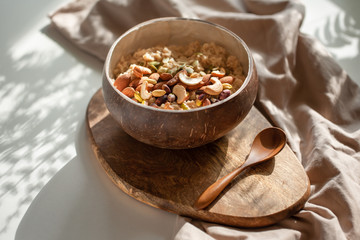 Oatmeal porridge in coconut bowl with wooden spoon on natural wooden tray. Porridge oats with almonds, pistachios, cashew and other nuts. Shadow of sunrise morning. White background. Side view.