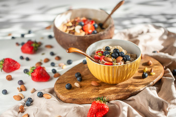 Oatmeal porridge in yellow dish with fruits and nuts on natural wooden tray. Porridge oats with strawberry, pistachios, blueberry, almond and banana. Shadow of sunrise morning. Side view