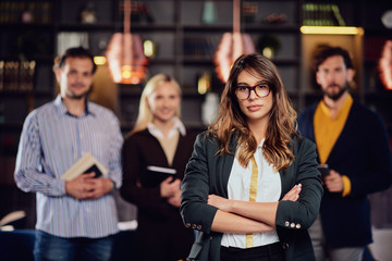 Serious Caucasian businesswoman with long brown hait and dressed smart casual standing in restaurand with crossed arms. In background her successful team posing.