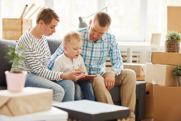 Content curious blond-haired son sitting with parents on sofa in living room with stacks of boxes and reading book on tablet while doing home task.