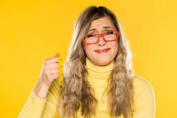 Dissapointed blonde with glasses, holding a measuring tape on yellow background