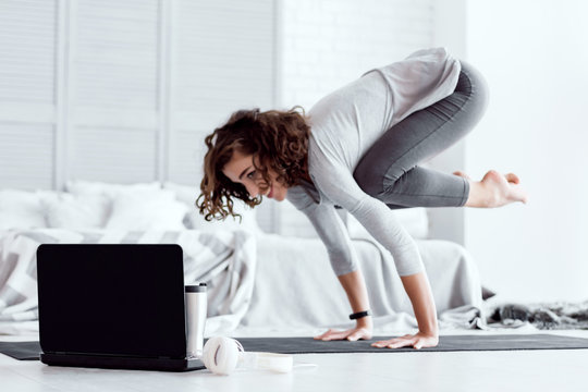 Young happy healthy fitness beautiful woman female doing bakasana yoga posture and workout on yoga mat opposite laptop with online master class at modern bedroom at home. Healthy lifestyle concept