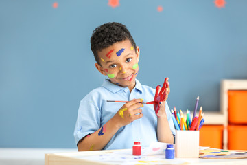 Cute African-American boy with hands and face in paint at home