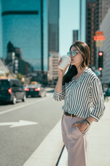 Professional young urban casual business woman in los angeles city out of concert hall standing on street having hot cup coffee waiting for yellow cab taxi. asian office lady drinking tea for morning