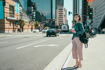 full length asian businesswoman in elegant wear with backpack standing on road holding coffee cup looking for taxi. confident female entrepreneur waiting for yellow cab on big city hurrying to office