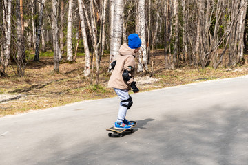 The boy rides a skateboard in the park in early spring. View from the back