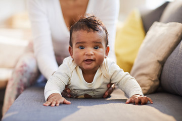 Portrait of positive frowning funny baby boy in bodysuit crawling on sofa while his mother supporting him