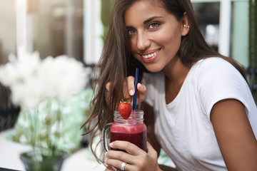 Close-up attractive coquettish tanned european young woman sip strawberry smoothie hold glass drink...