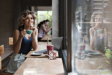 Dreamy happy lucky young alluring girl sitting coffee table cafe drink cappuccino, smoothie gazing through window passersby thoughtful creating project working freelance, digital nomad use laptop