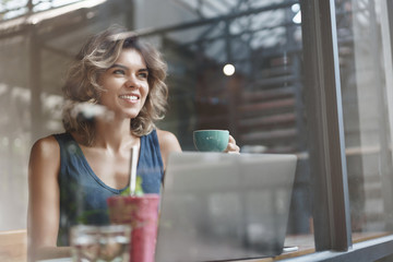 Charismatic charming european blond curly-haired successful woman drink coffee enjoy lunch sit cafe window bar look outside delighted taste coffee hold cup use laptop, researching, working freelance