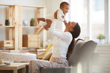 Jolly excited young African woman with pony tail enjoying maternity and having fun with baby son at...
