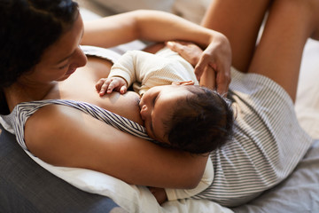 Close-up of serene loving young black mother in nightgown sitting on bed and breastfeeding baby in...