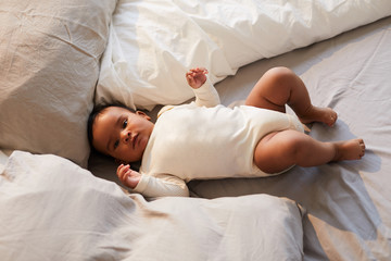 Portrait of serious adorable African baby boy in bodysuit lying between pillows in comfortable bed