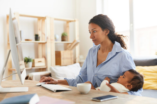 Serious concentrated young African mother designer in oversize shirt sitting at desk and working on project while nursing baby