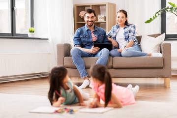 family, leisure and people concept - happy mother and father with tablet pc computer sitting on sofa and looking at daughters drawing at home