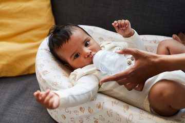 Close-up of serious frowning cute little black boy in bodysuit lying in cocoon and drinking clear water from baby bottle given by mother