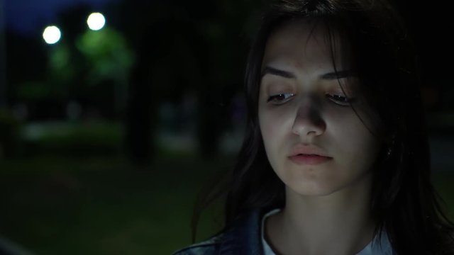 beautiful pensive caucasian girl using smartphone and device for smoking electronic tobacco heating system on the street at night