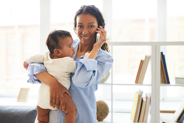 Positive confident modern young African mom standing against shelves and holding baby in arm while talking on mobile phone and looking at camera
