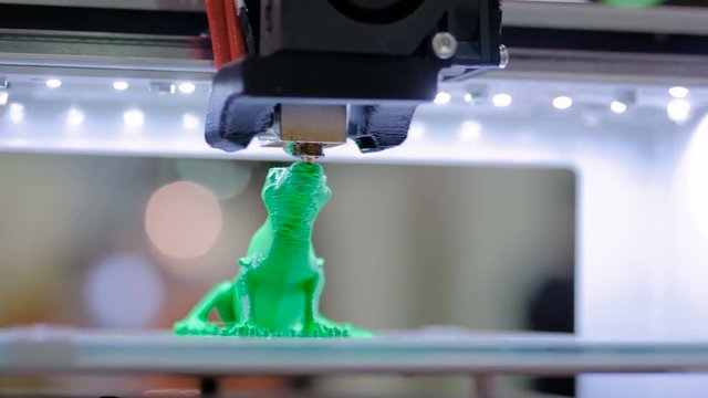 Close up shot - 3D printer prints physical 3D model of green toy lizard with plastic wire filament at modern technology exhibition. Additive technologies, science and futuristic concept