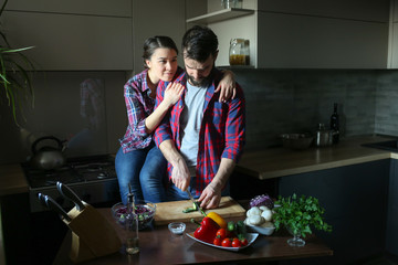 Beautiful young couple in kitchen at home while cooking healthy food. Man is cuts salad. Woman sits on table and hugs man from behind. Scene from family life.