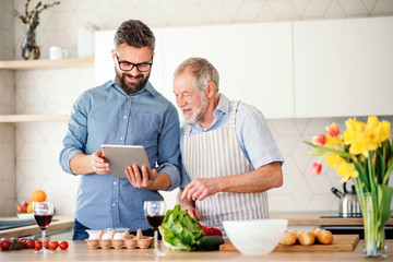 Adult hipster son and senior father indoors in kitchen at home, using tablet.