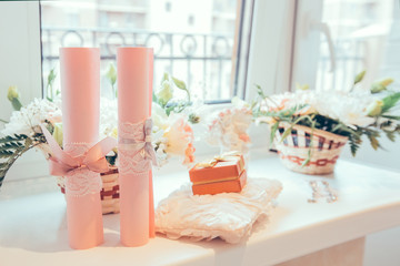 Wedding accessories near the window in bedroom of bride: two pink scrolls with lace, wedding rings, flowers, pillow for rings, jewerly box. Romantic greeting card, engagement, celebration, invitation