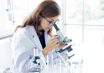 Female scientist in white coat doing a research using microscope in laboratory. Long hair asian woman wearing eyeglasses.