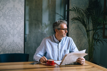 middle aged gray haired man reading newspaper and drinking coffee in cafe bar