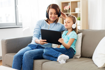 people, family and technology concept - happy mother and daughter with tablet pc computer and headphones listening to music at home