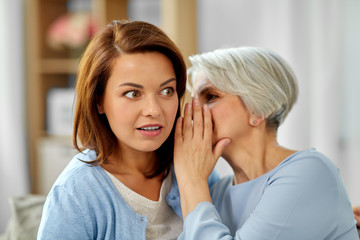gossiping and family concept - senior mother whispering secret to adult daughter at home