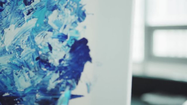 Dolly zoom in closeup shot of blue and white abstract acrylic painting being created on canvas with palette knife by unrecognizable female artist