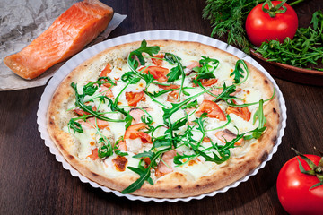 Top view on pizza with tomato, parmesan and salmon with white sauce with ingridients on brown wooden table. Picture for recipe or menu. Copy space for design. Served and cooked pizza. Italian cuisine