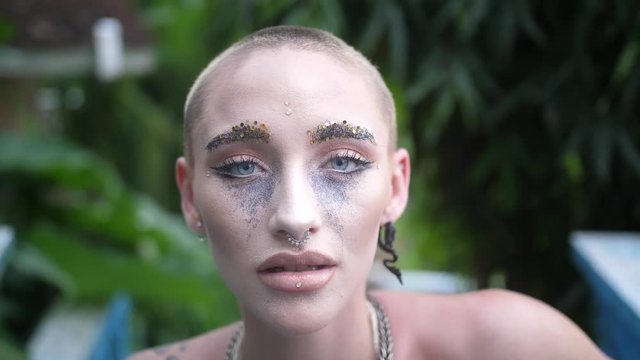 Woman with leopard print tattoo and shaved head dancing outside in a tropical garden. Lady with glitter makeup on her eyes and face moves slowly while looking into camera.