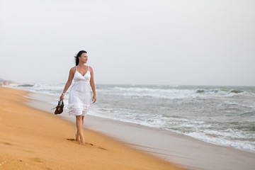 beautiful young woman in white dress walking on the sandy beach, holding sandals. travel and Summer concept