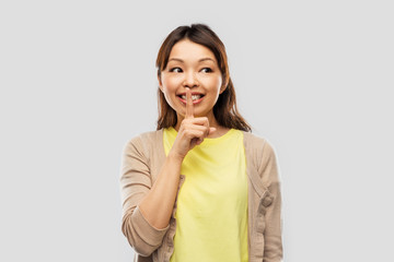 people, silence and secret concept - happy asian young woman with finger on lips over grey background