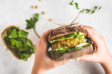 Vegan sandwich with chickpea patty, avocado, cucumber and greens in rye bread in children's hands. - obrazy, fototapety, plakaty