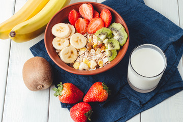 Cereal muesli with strawberries, banana and kiwi in the bowl, glass with milk on wooden white background, healthy breakfast