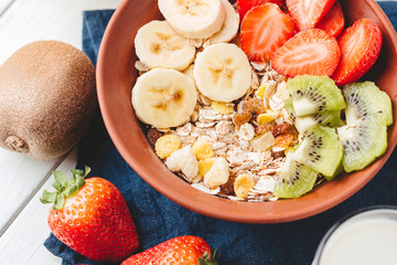Cereal muesli with strawberries, banana and kiwi in the bowl, glass with milk on wooden white background, healthy breakfast